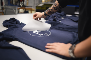 Image de production d'un transfert textile DTF haute définition sur un sweat-shirt bleu marine. Une main  décolle le film protecteur après pressage, révélant un logo blanc net et complexe. L'arrière-plan montre un atelier d'impression moderne.